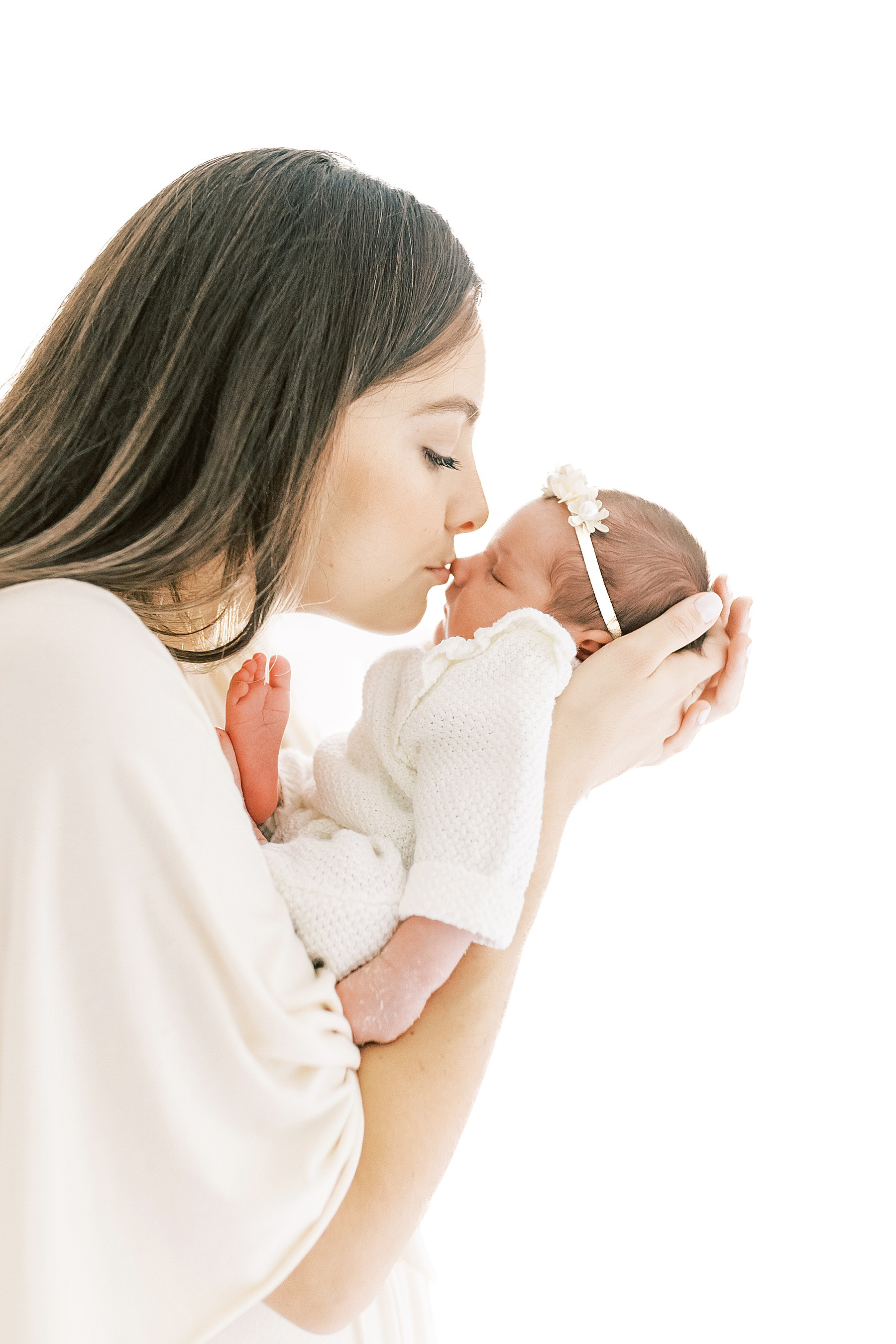 woman in cream long dress standing against white sheer curtains kissing newborn baby girl on the nose