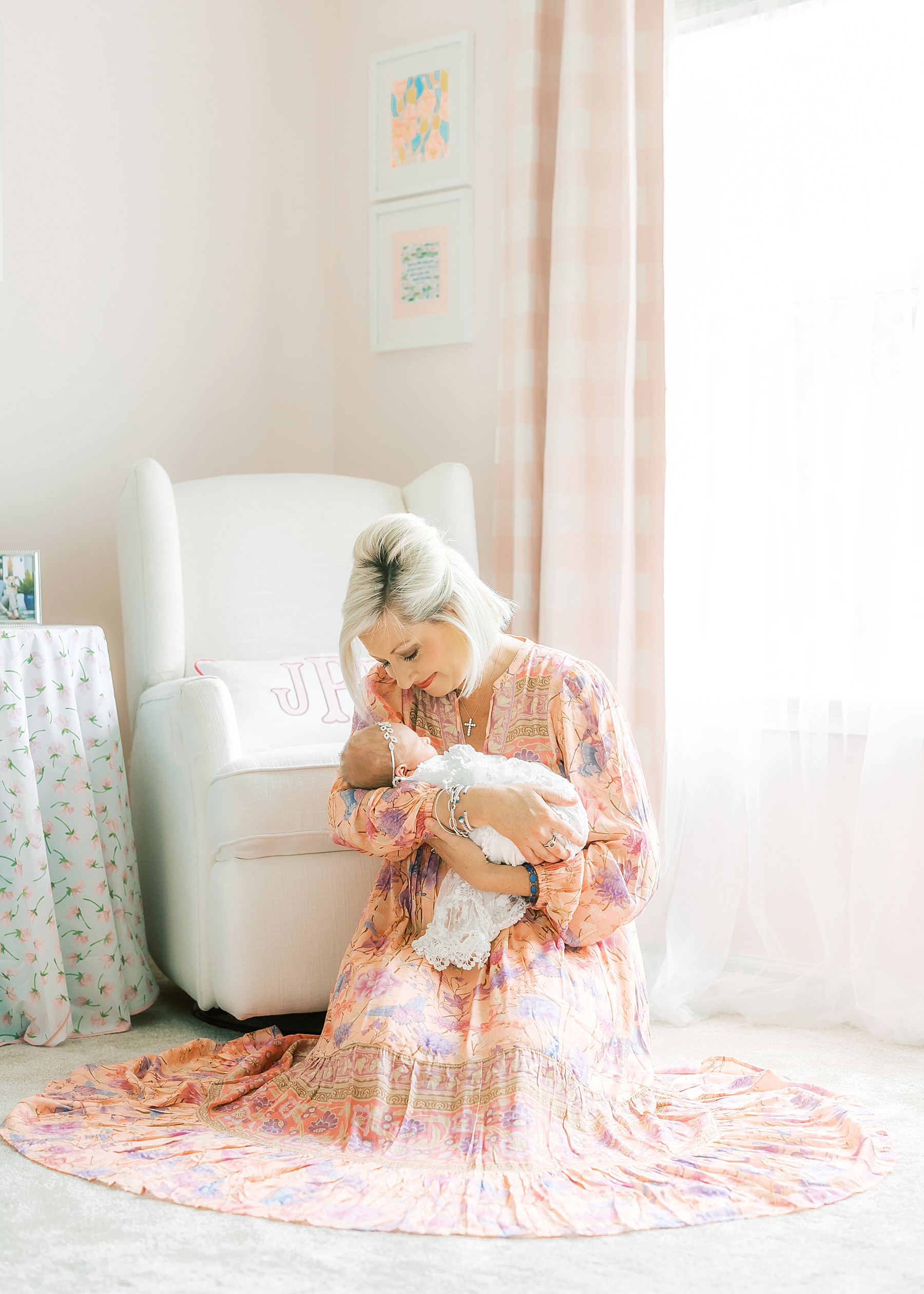 woman holding a newborn baby girl wrapped in white lace wearing pink floral boho maxi dress, woman sitting down on floor in baby girl nursery, pink walls