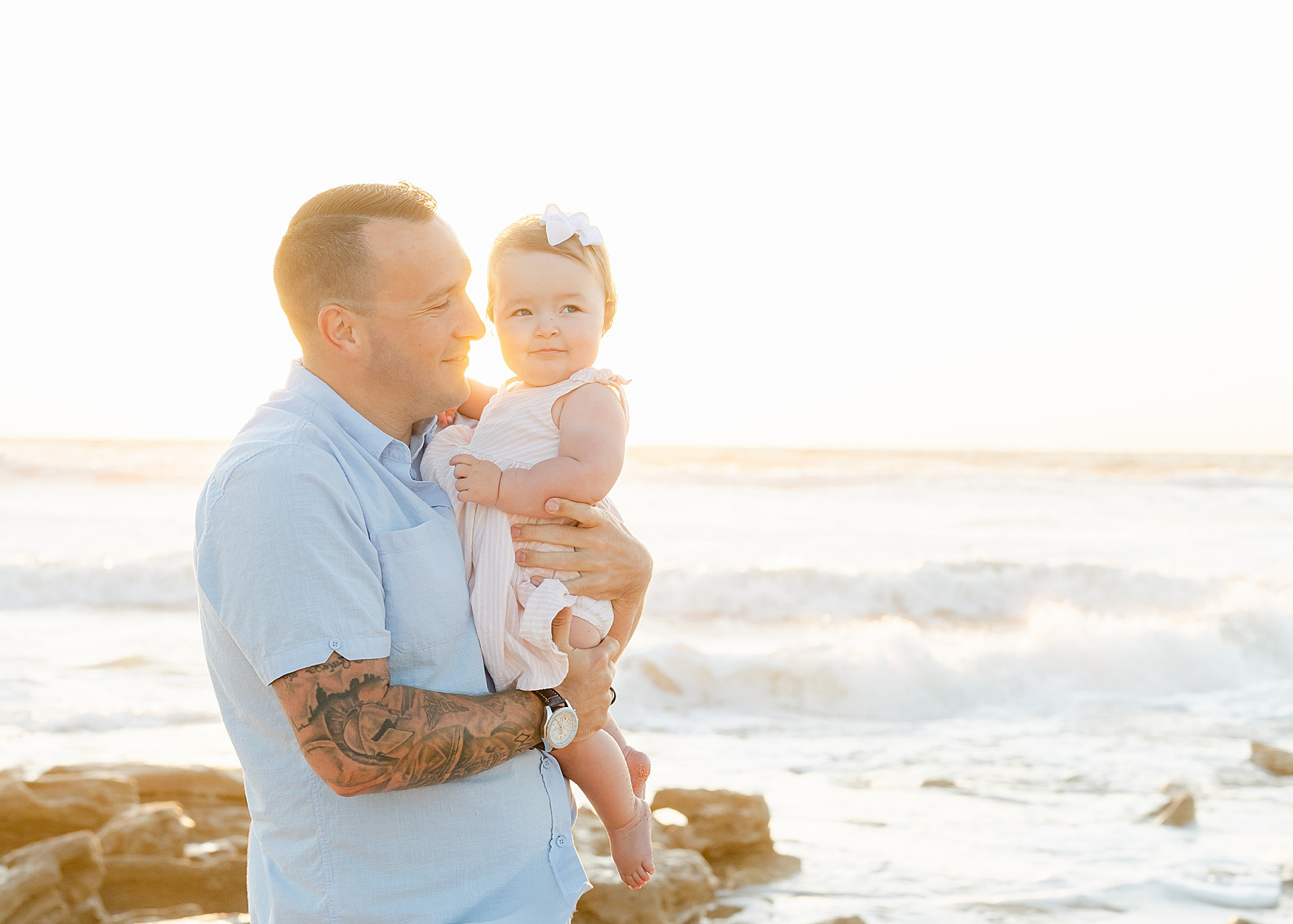 man in light blue shirt holding baby girl on the beach at sunrise