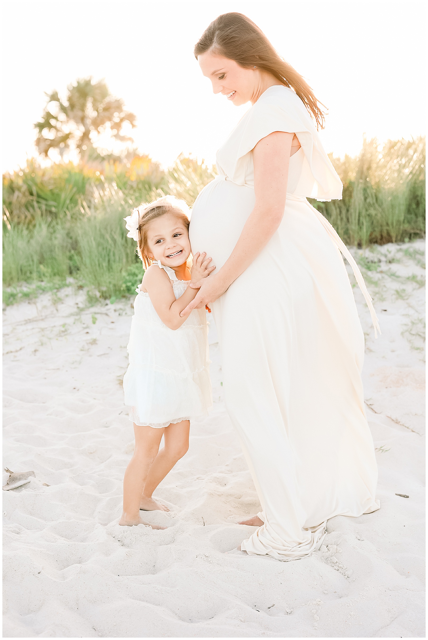 pregnant woman in cream maxi dress holding little girl on the sand on the beach