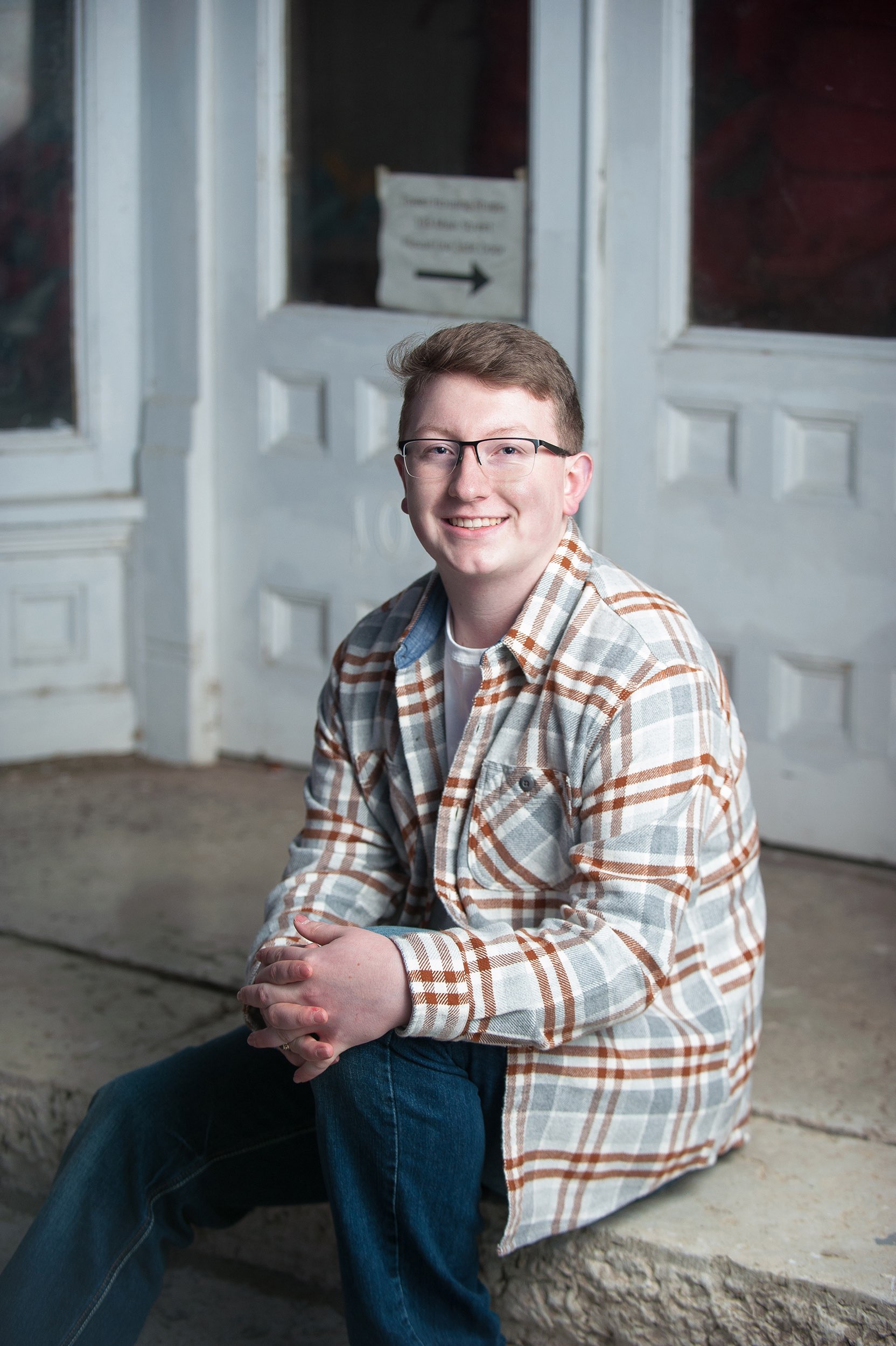 Senior guy in plaid shirt sitting in front of a white building in Galena, MO.