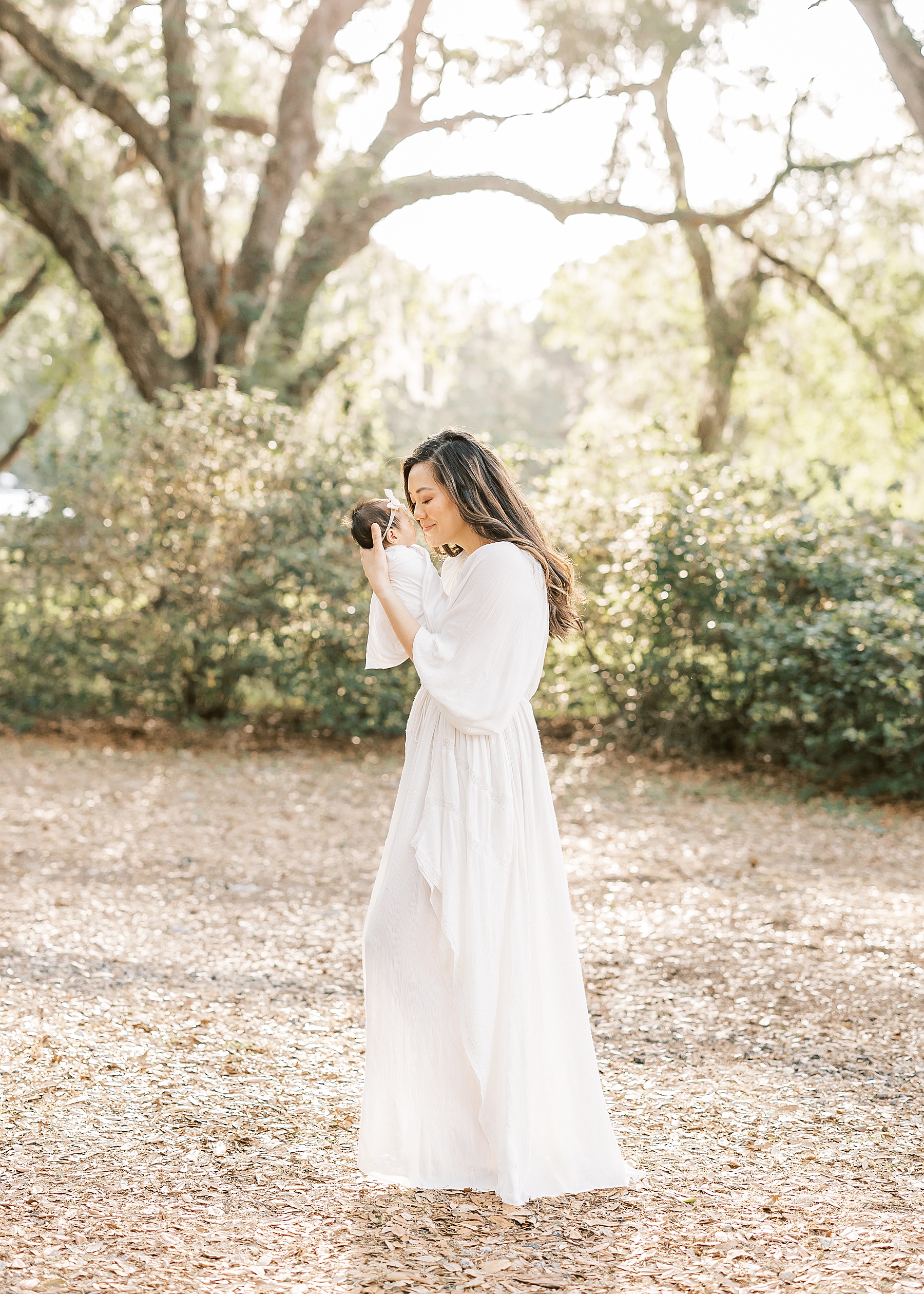 sun haze filled image of woman in long white dress holding newborn baby girl wrapped in white swaddle