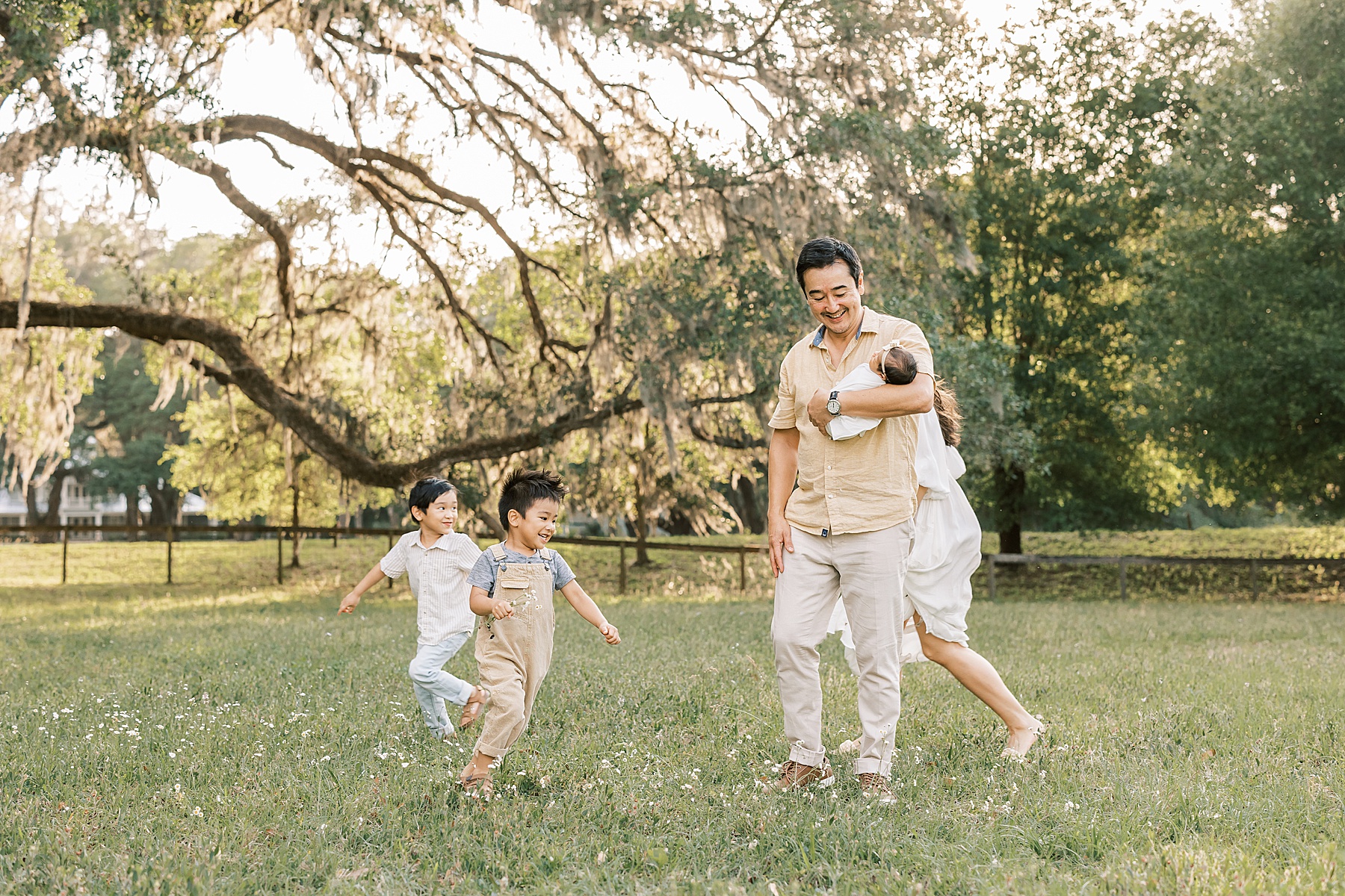 man holding newborn baby girl in sunny filed in the spring with children running around him