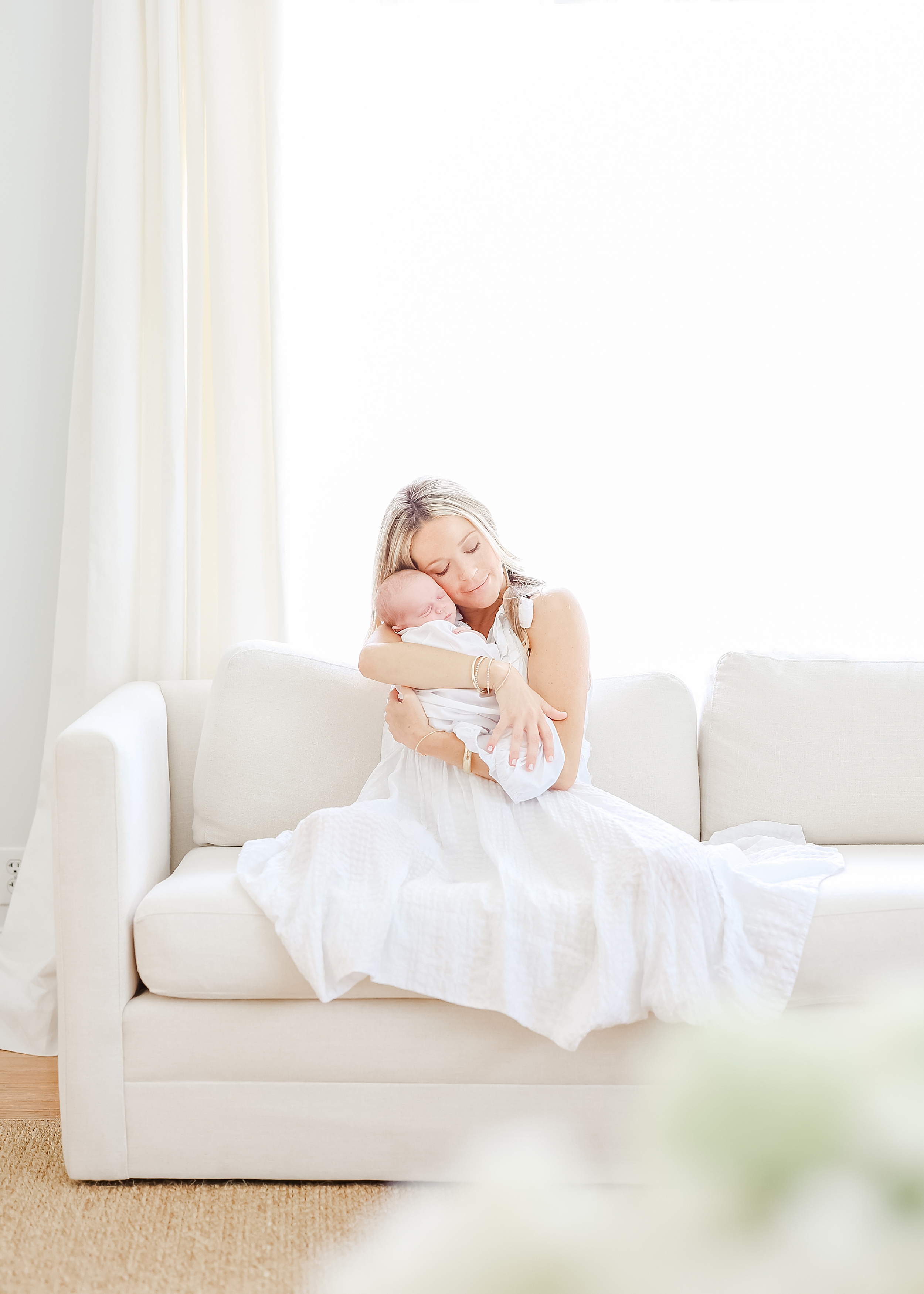 Woman dresses in long white dress holding newborn baby boy on white couch in front of window with sheer airy sunlight.