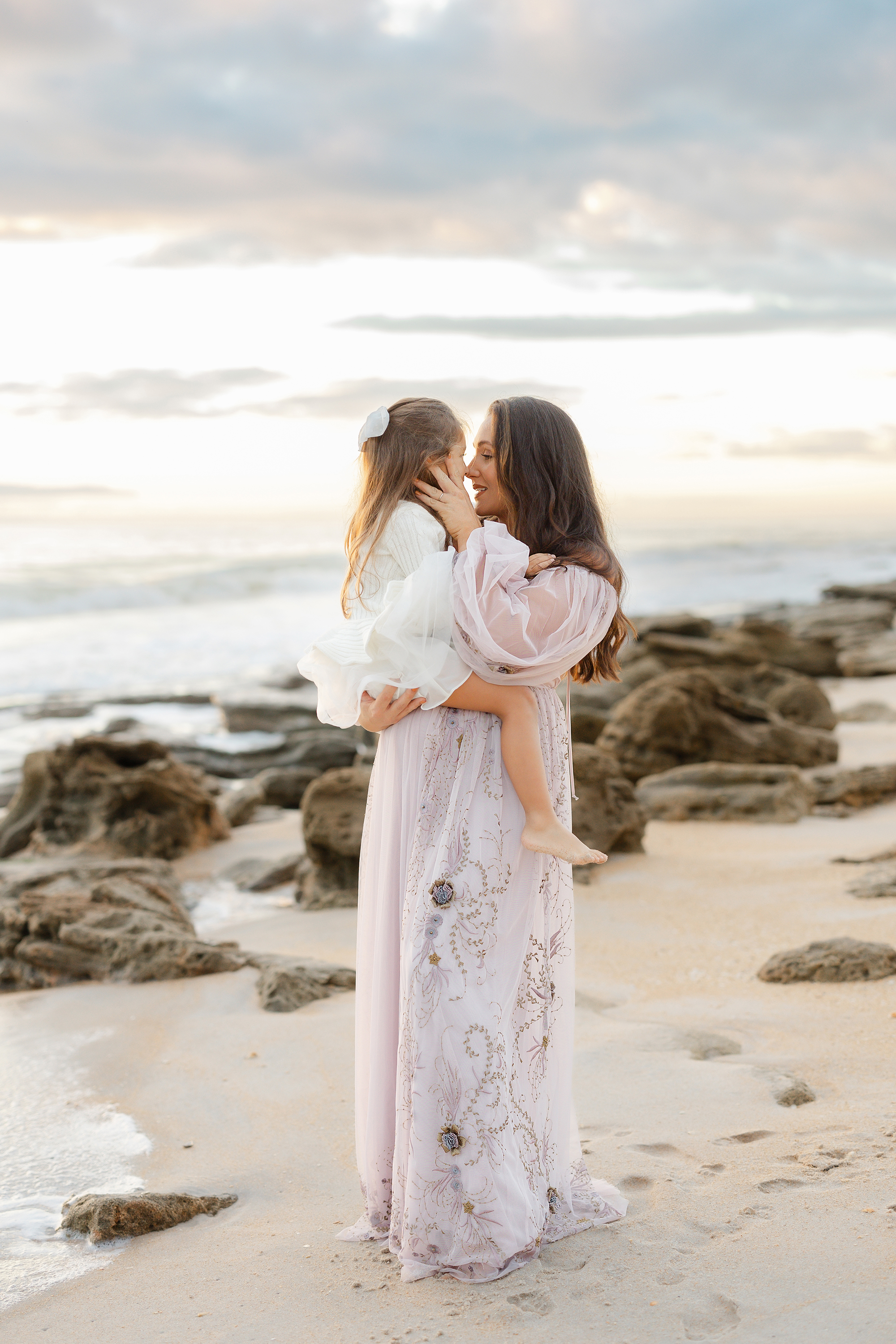 Woman holding her little girl in neutral colors on the beach at sunrise.