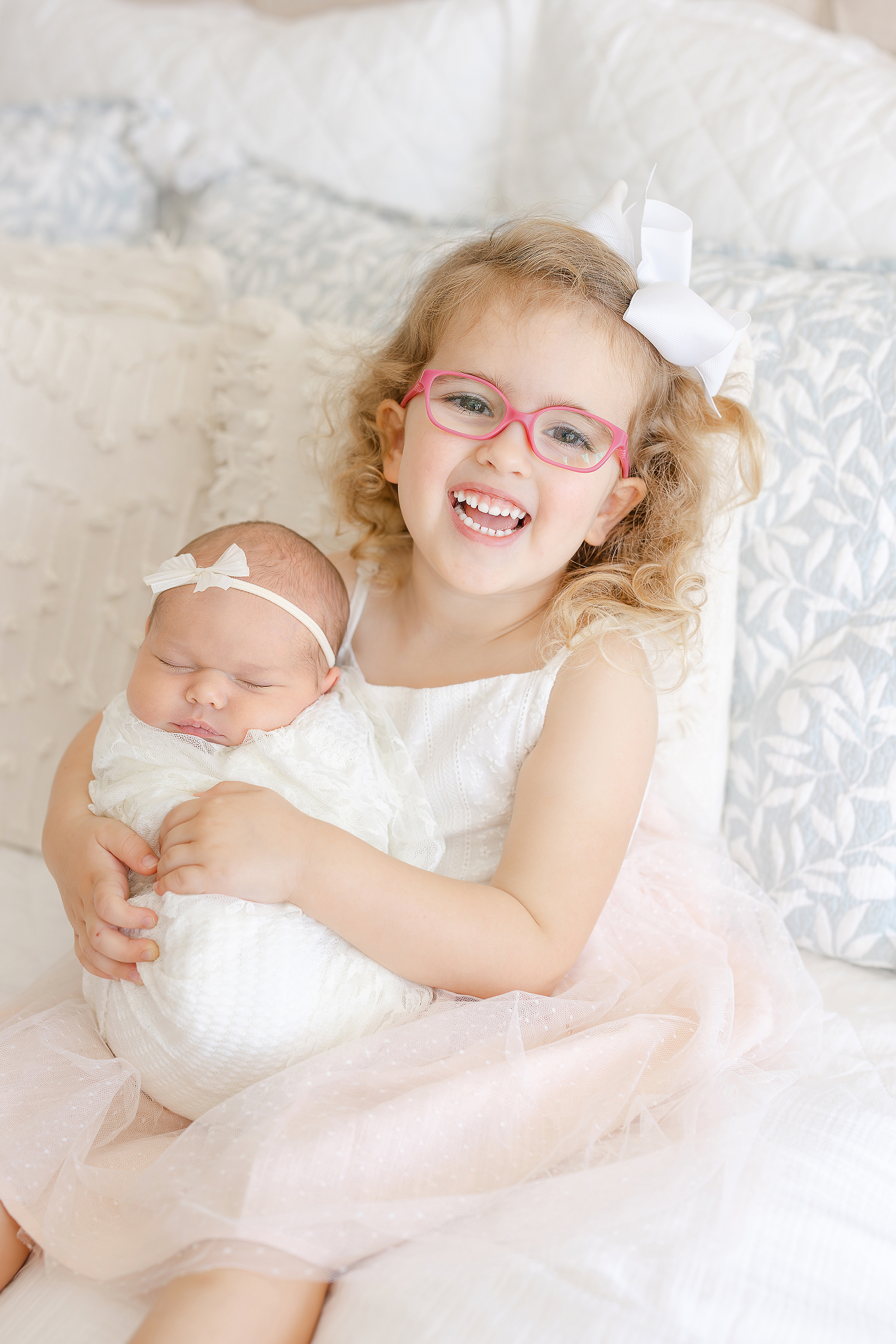 A sibling portrait of a little girl in hot pink glasses holding her newborn baby sister.