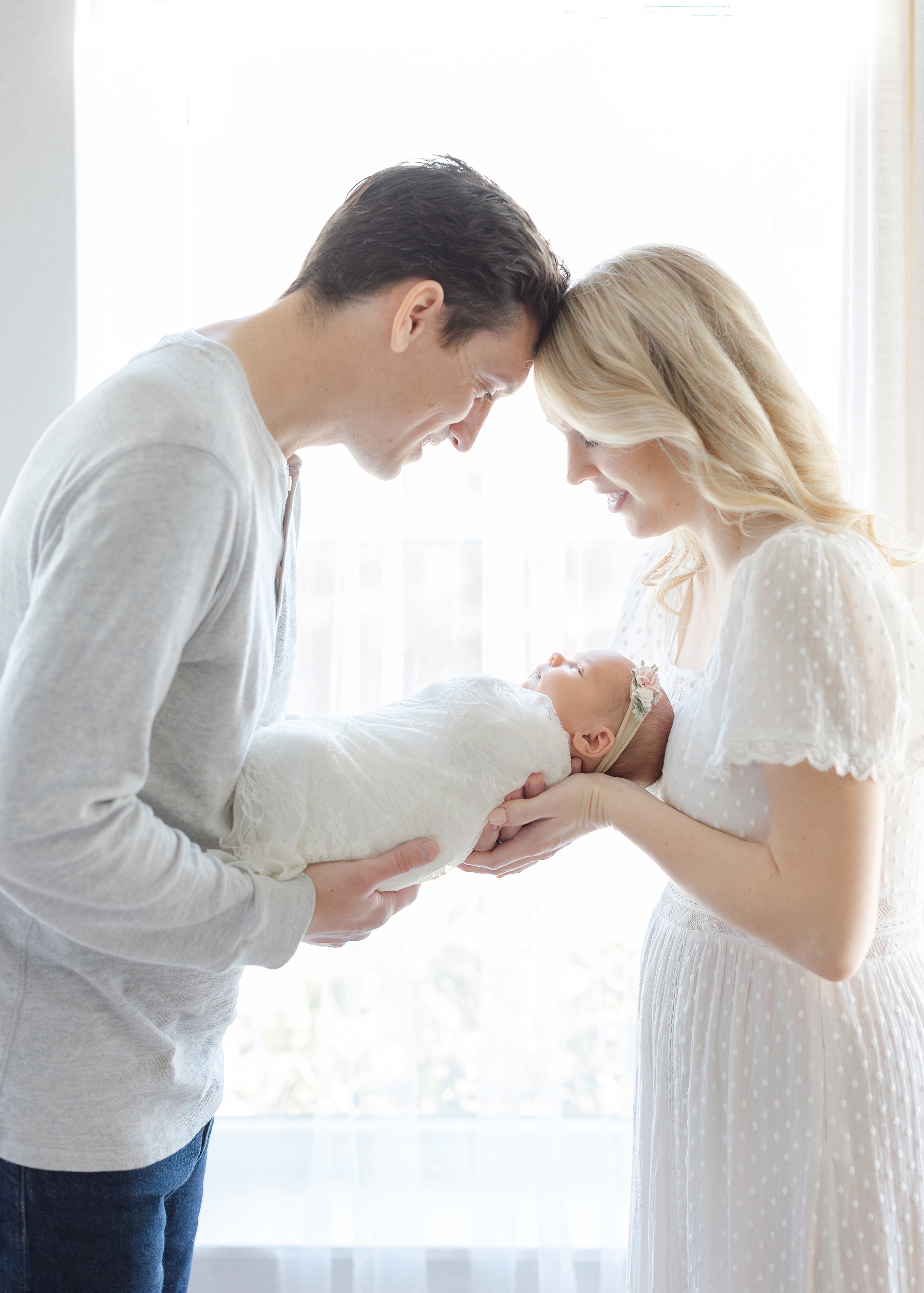 A light and airy portrait of a couple holding their newborn baby girl in front of a window.