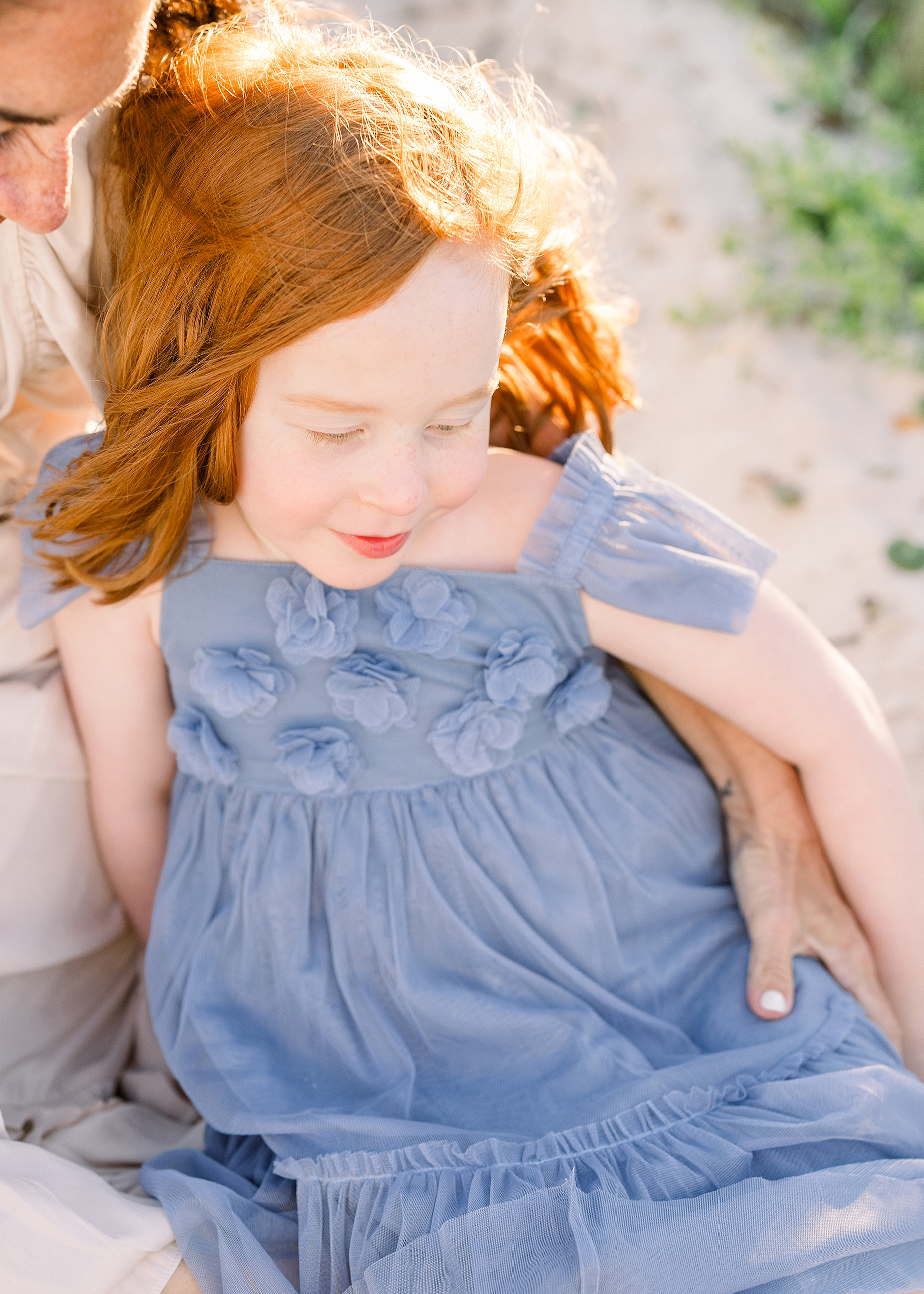 A little girl with red hair in a purple dress sits on the sand at the beach at sunset.