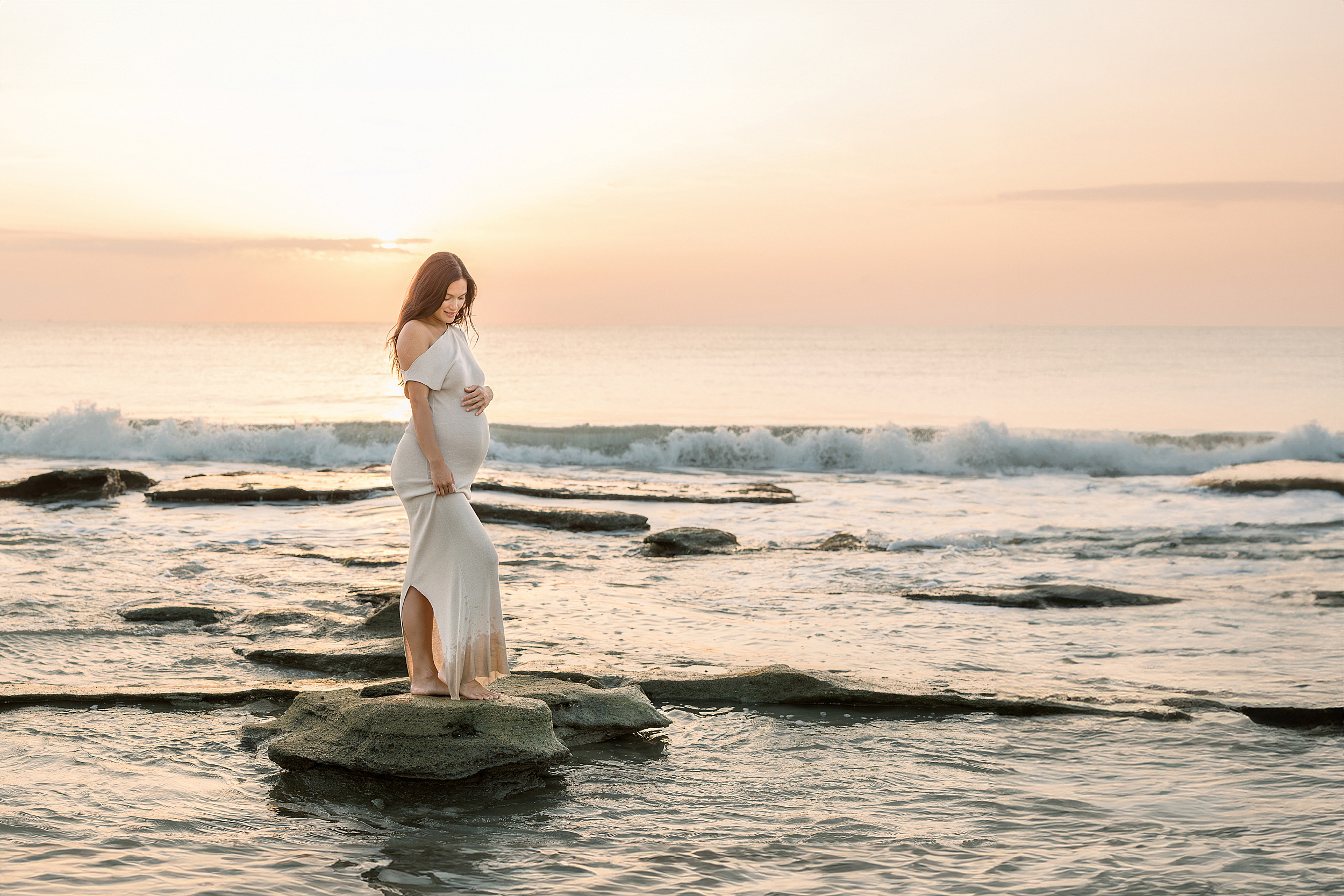 A sunrise pregnancy portrait of a woman standing on the rocks at the beach in Saint Augustine, Florida.