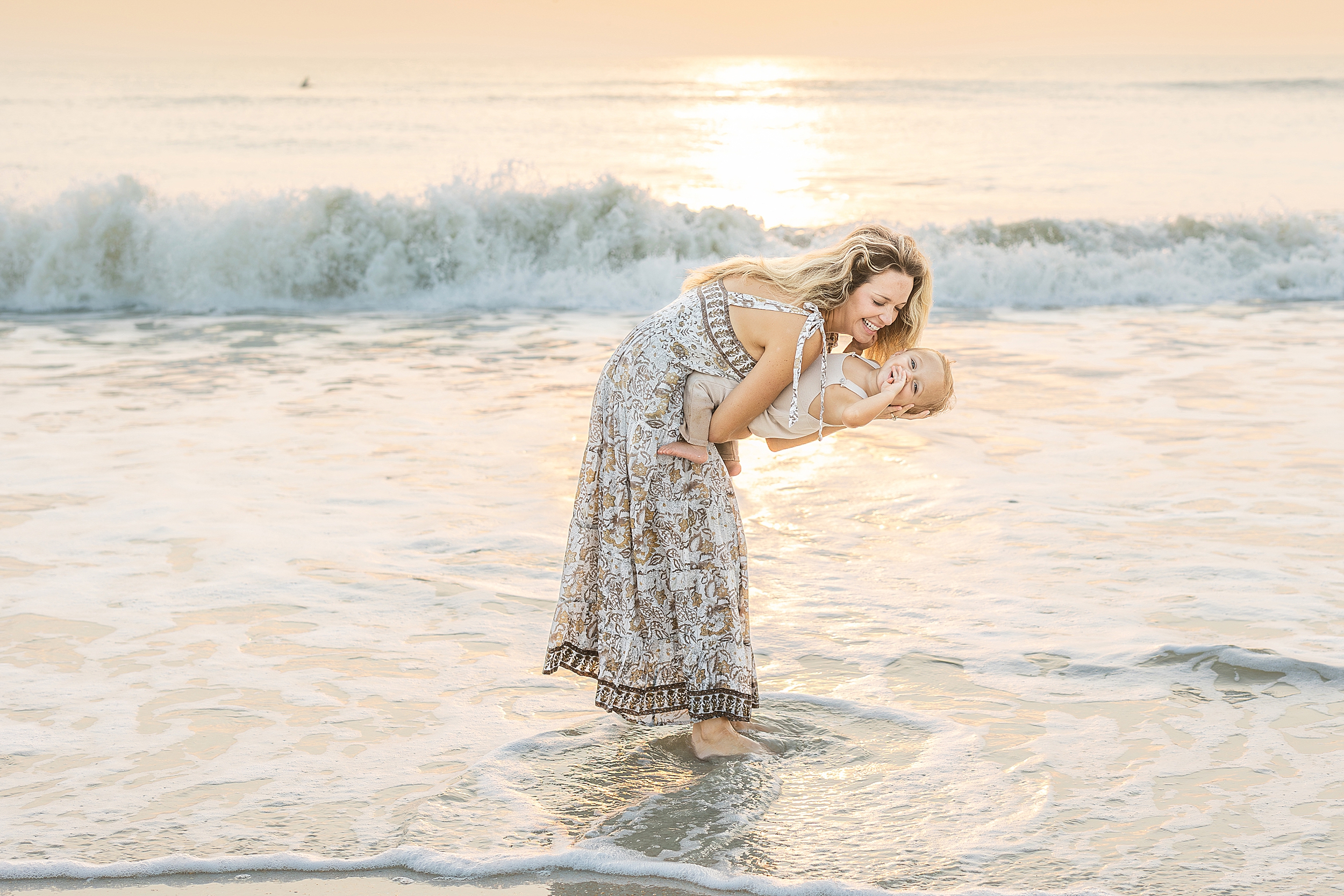 A woman stands in the water at the beach at sunrise and dips her baby boy in the sunlight.