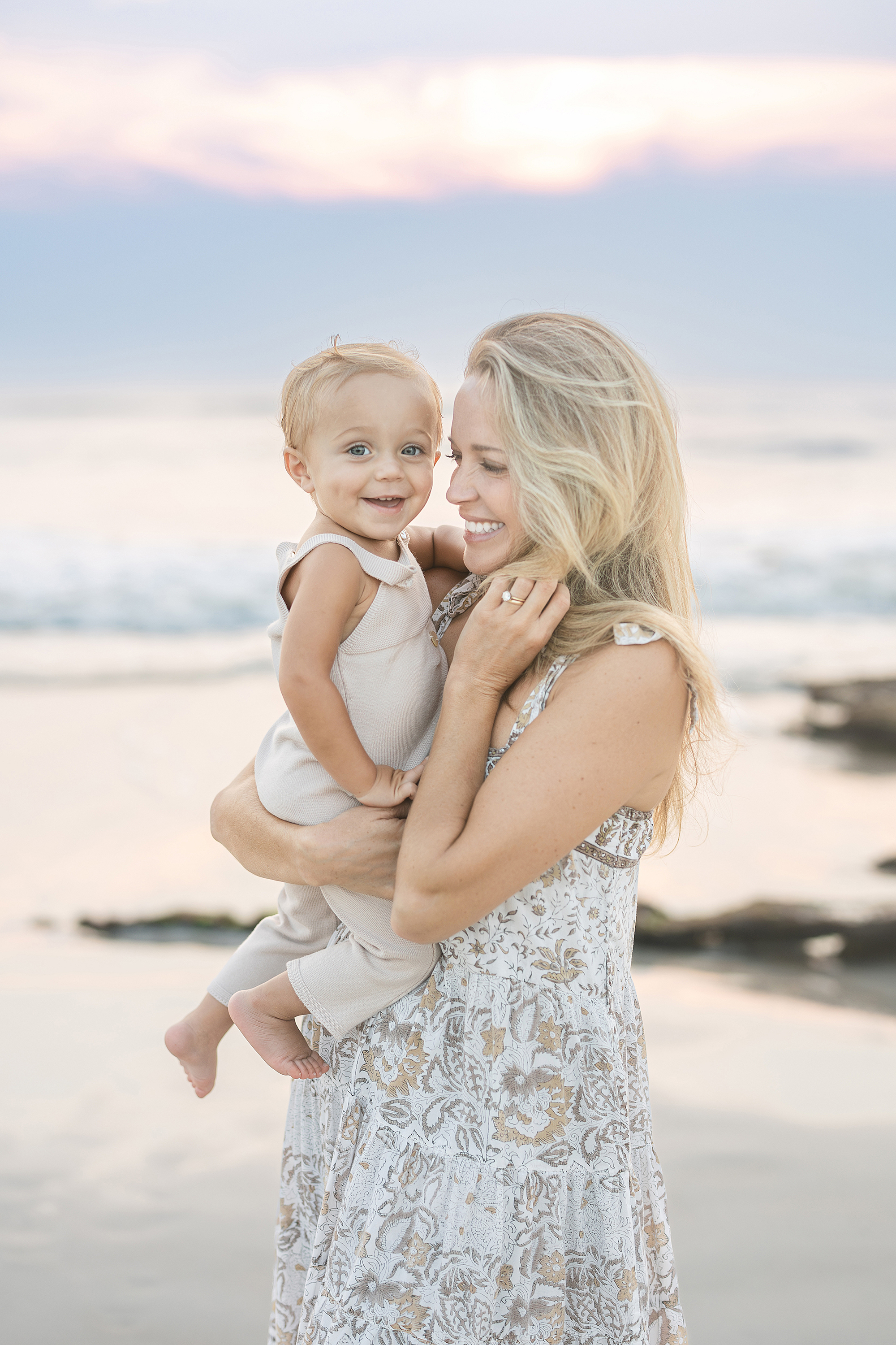 A mother holds her smiling one year old baby boy on the beach at sunrise. 