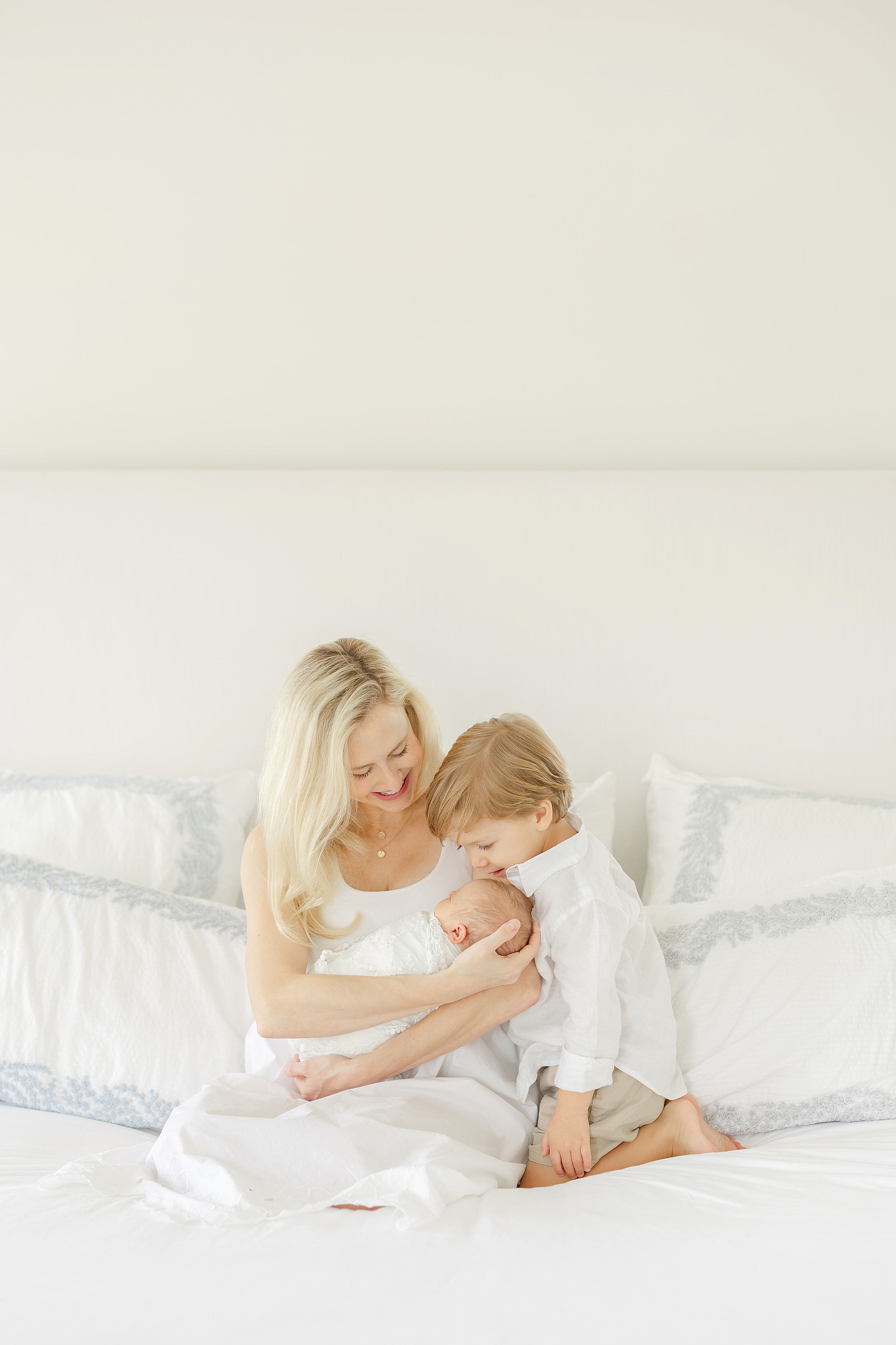 An airy newborn portrait of a mother in a white dress on her white bed with her two young children.