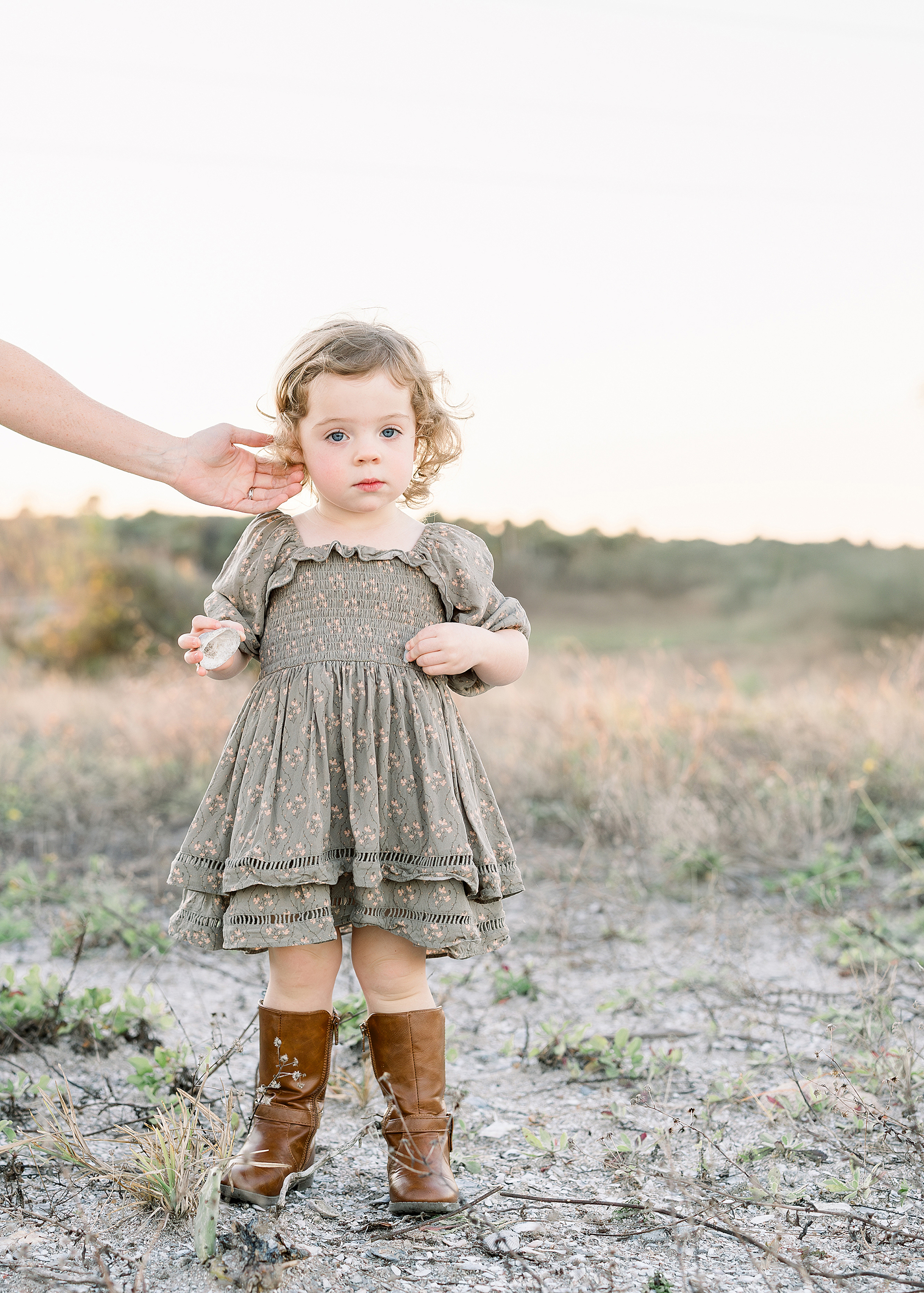 A portrait of a little girl in a neutral green dress stares at the camera while her mother brushes her hair from her face.