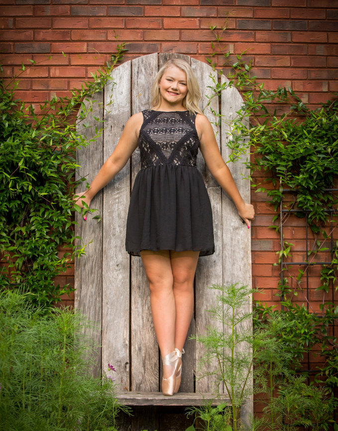 Woman in black dress poses in ballet shoes against a rustic wooden background, surrounded by green plants.