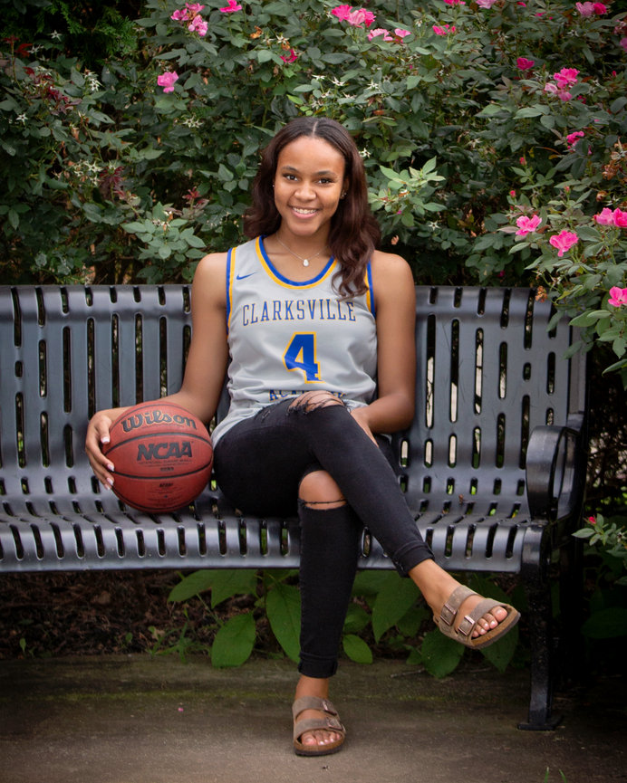 A woman in a basketball jersey sits on a bench with a ball, surrounded by pink flowers.