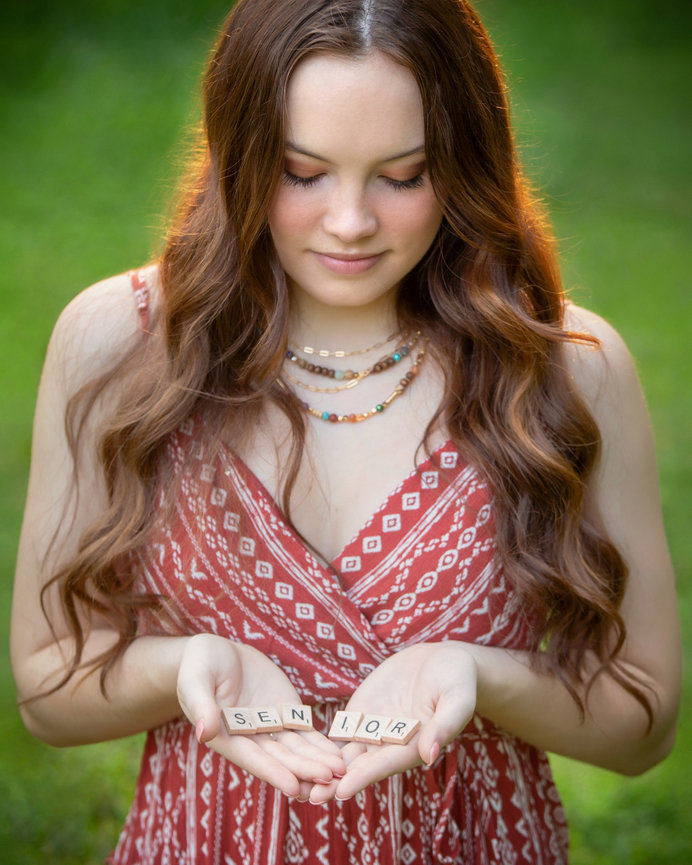 Young woman in a patterned dress holds scrabble tiles spelling SENIOR outdoors.