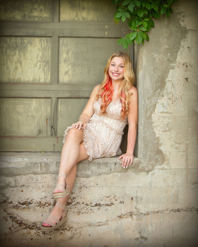 Woman with long hair sitting on ledge in a patterned dress, near an old building with green vines, smiling.