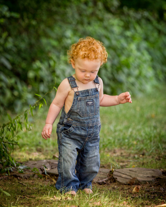 Toddler with curly red hair in denim overalls walking on grass outside.