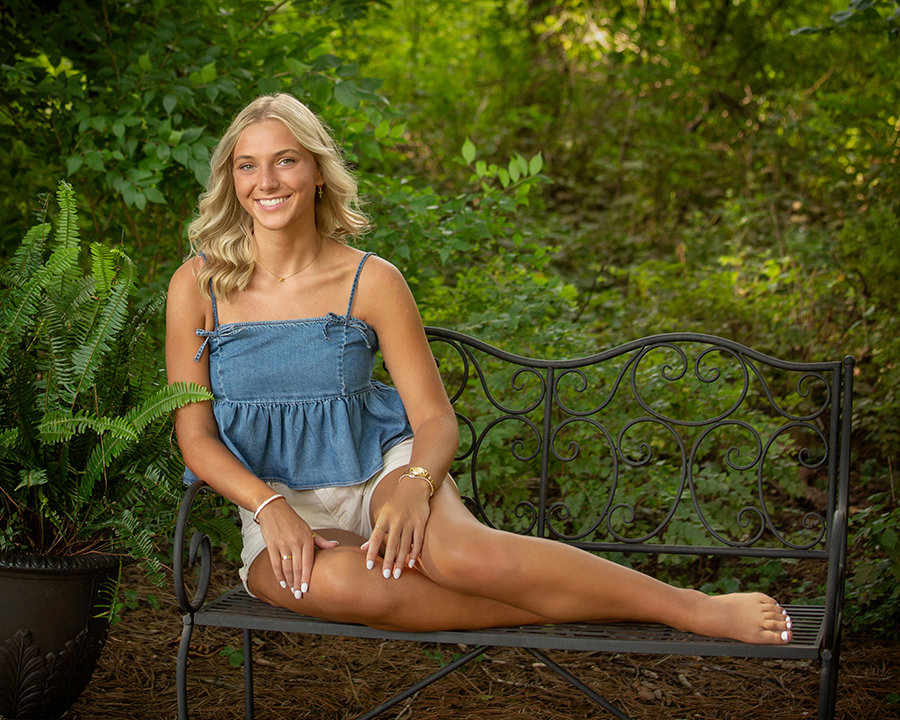 A person with blonde hair sits on a wrought iron bench outdoors, surrounded by greenery, wearing a blue top and shorts.