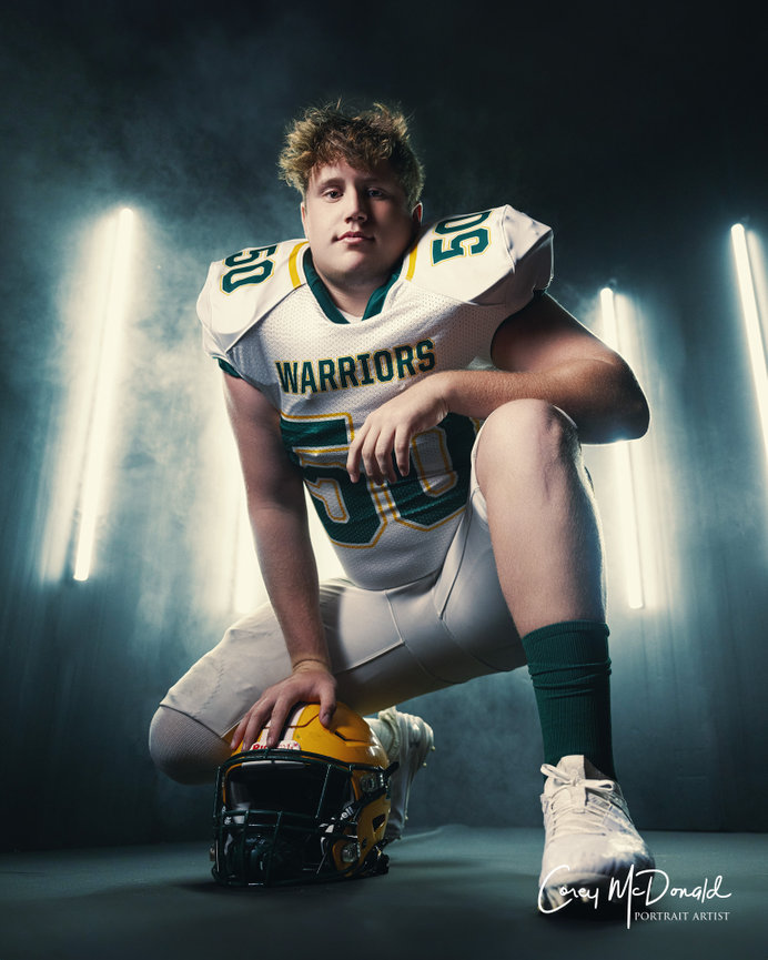 Football player kneeling in uniform and holding helmet, with bright lights in the background.