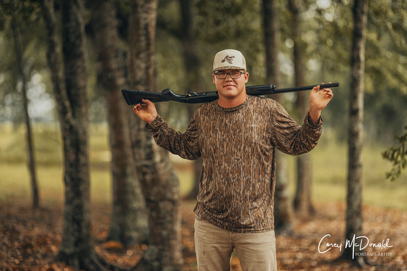 Man in camouflage shirt holding a rifle on his shoulders, standing in a forested area.