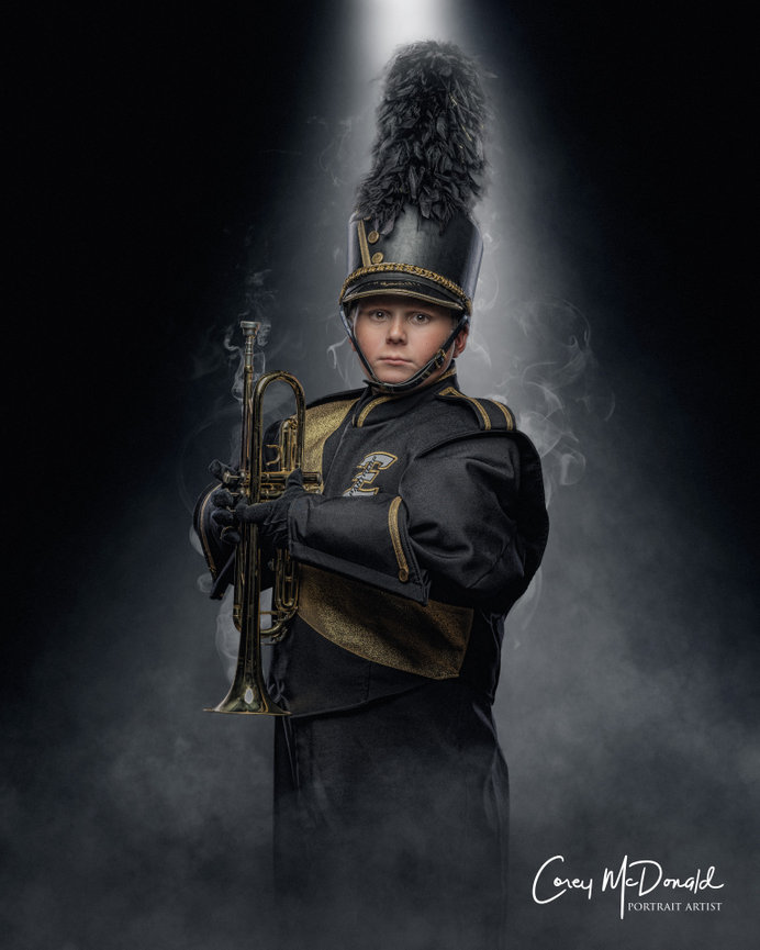 A marching band member in uniform holding a trumpet, illuminated by spotlight, with a smoky background.