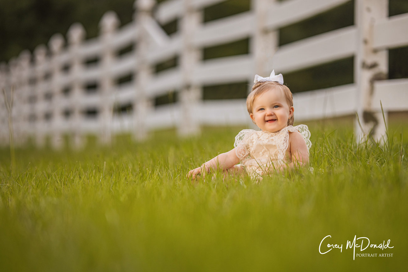A smiling baby in a lace dress sits on grass near a white fence.
