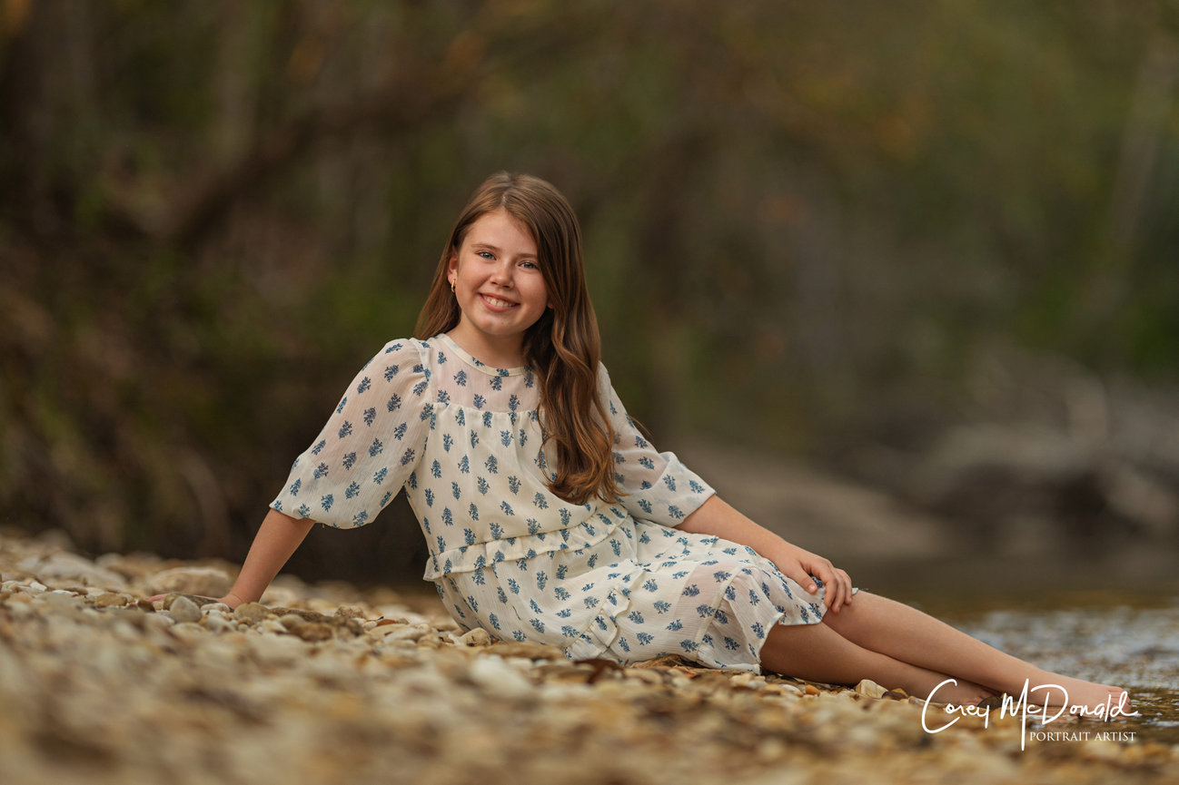 Girl in floral dress sitting on pebbles by a riverbank, smiling, with lush trees in the background.
