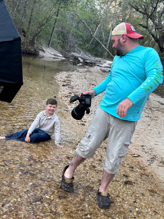 Man with camera stands by a stream looking at a boy sitting in the shallow water, surrounded by trees and nature.