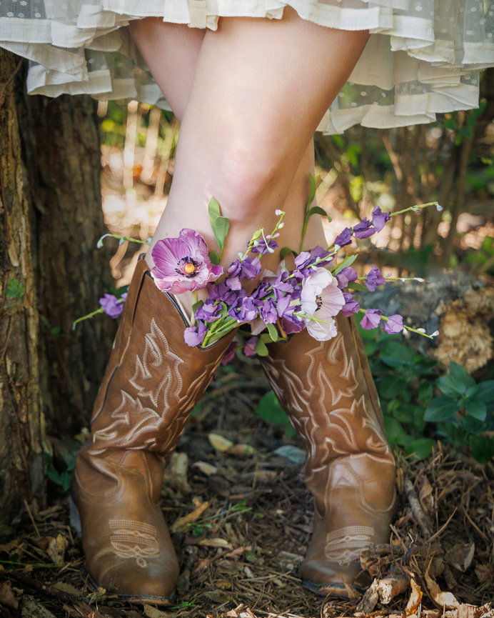 Cowboy boots filled with purple flowers, worn by a person in a white dress, standing outdoors on a leafy ground.