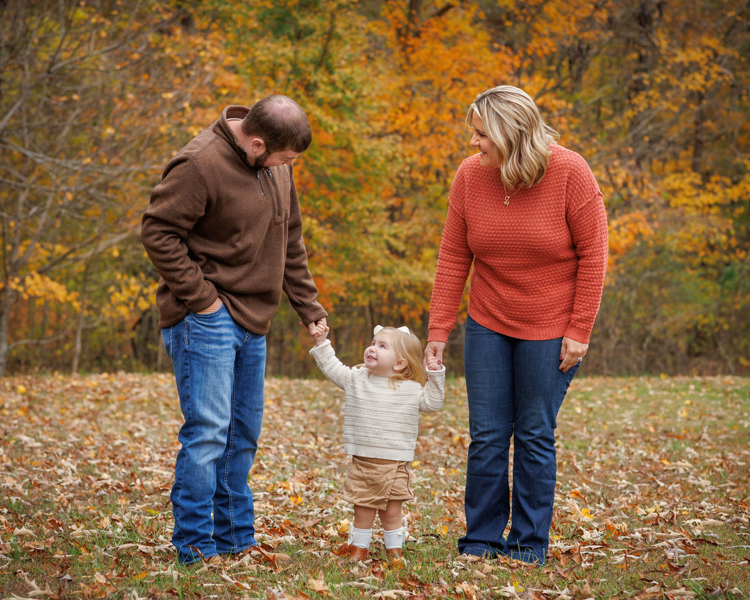 A family of three holding hands in a fall forest setting with colorful autumn leaves.