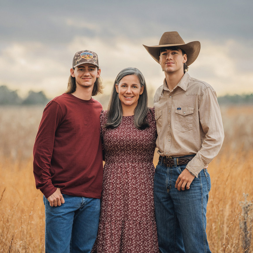 Three people stand in a field: a woman in a floral dress flanked by two men in jeans and hats, under a cloudy sky.