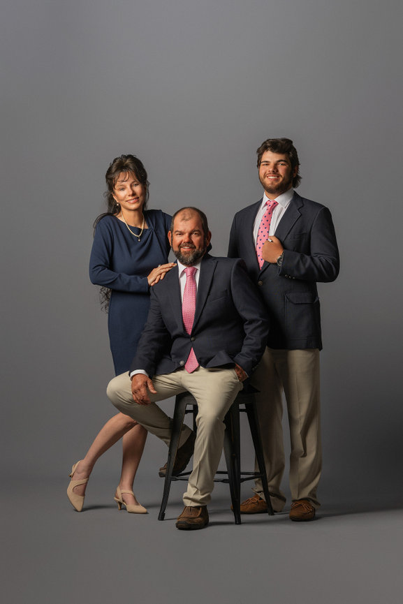 A woman and two men in formal attire pose against a grey background. The man is seated, while the others stand beside him.
