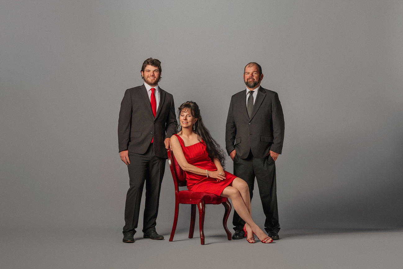 Three people in formal attire; two men in suits, one seated woman in a red dress, against a gray background.