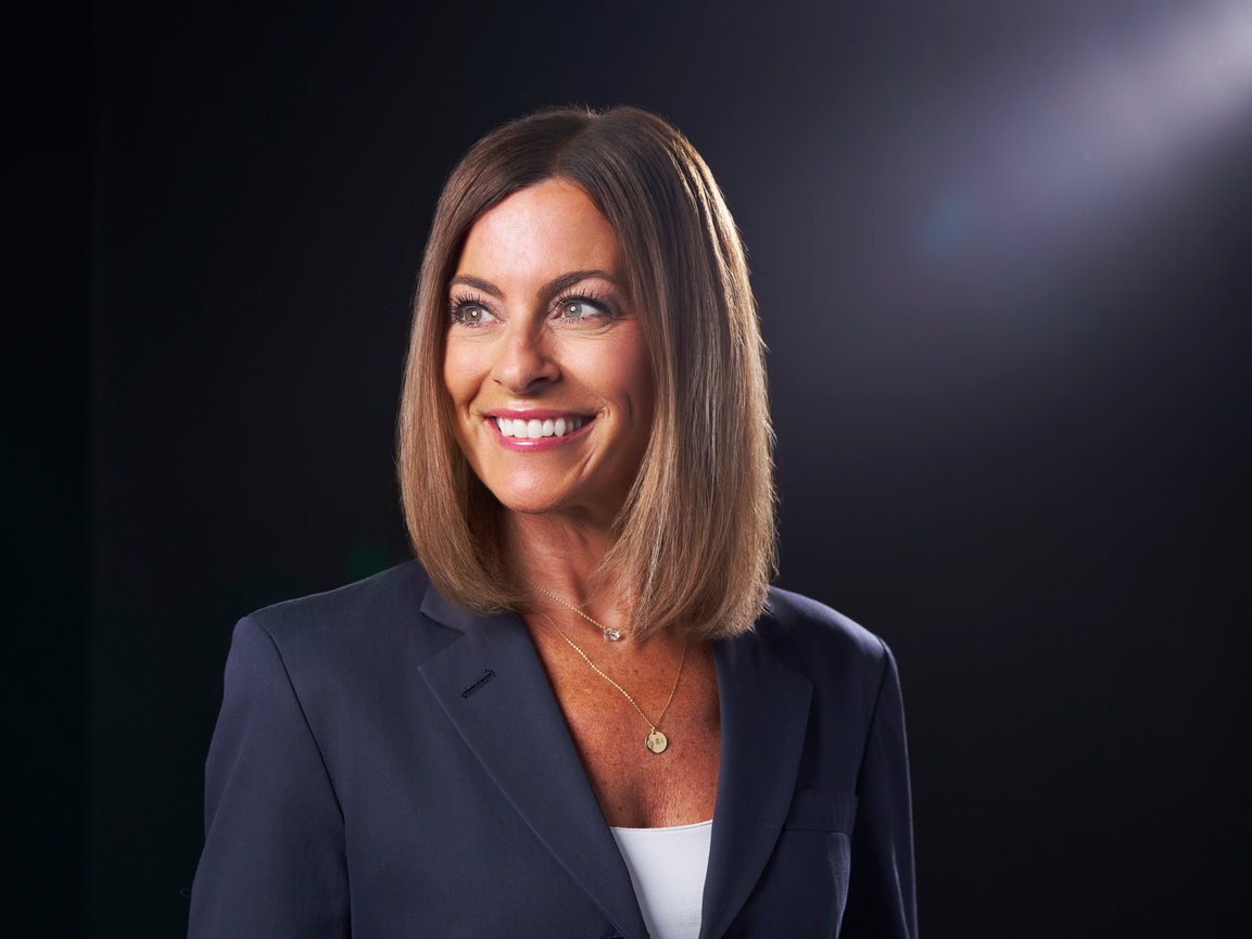 Portrait of a smiling woman in a dark blazer against a black background with soft light from the right