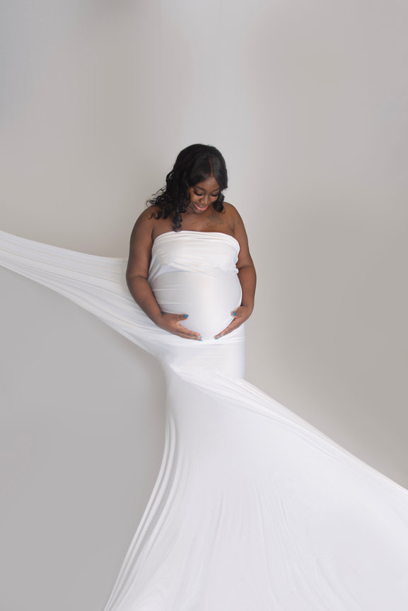 Pregnant woman in a white strapless gown, gently holding her belly with a flowing fabric backdrop.