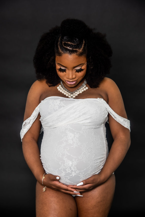 Pregnant person in a white lace outfit, smiling while cradling their belly, against a black backdrop.