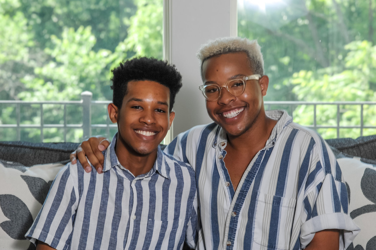 Two smiling young men sitting together on a couch, wearing matching striped shirts, with greenery visible in the background.