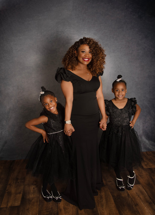 A smiling woman in a black outfit holding hands with two girls in black dresses, posing together.