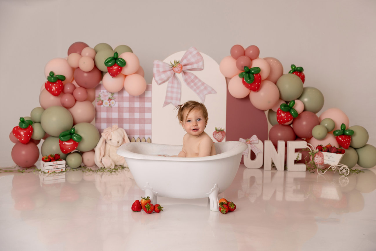 Toddler in a white tub at a strawberry-themed first birthday party with balloons and pastel decor