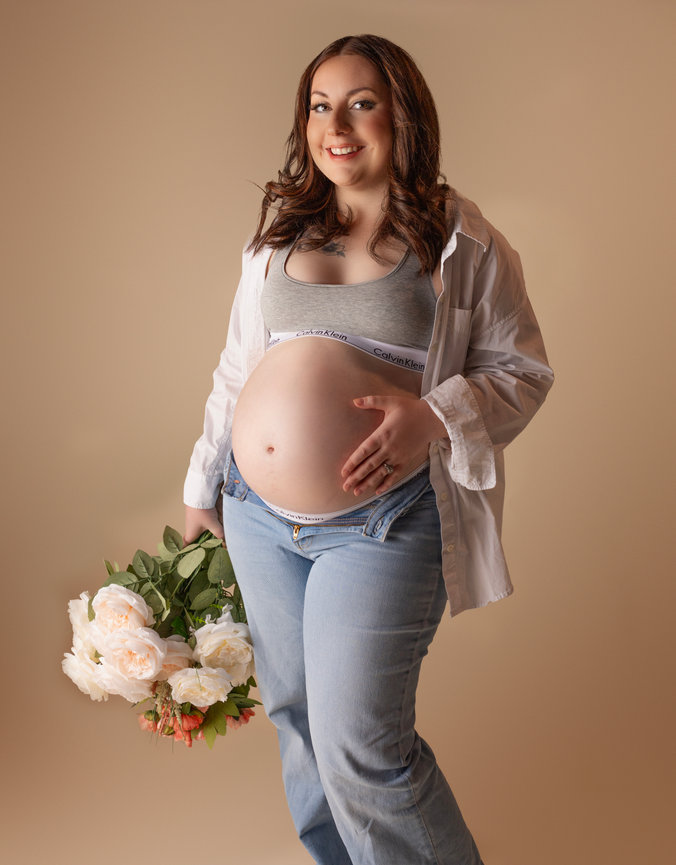 Modern maternity portrait of a glowing mom-to-be in jeans, bralette, and open white shirt. Holding soft florals, she radiates confidence and beauty in a warm studio. A luxury maternity photography experience for stylish mothers in Calgary & Okotoks