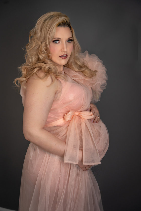 Pregnant woman in a flowing, pink dress posing against a dark gray background.