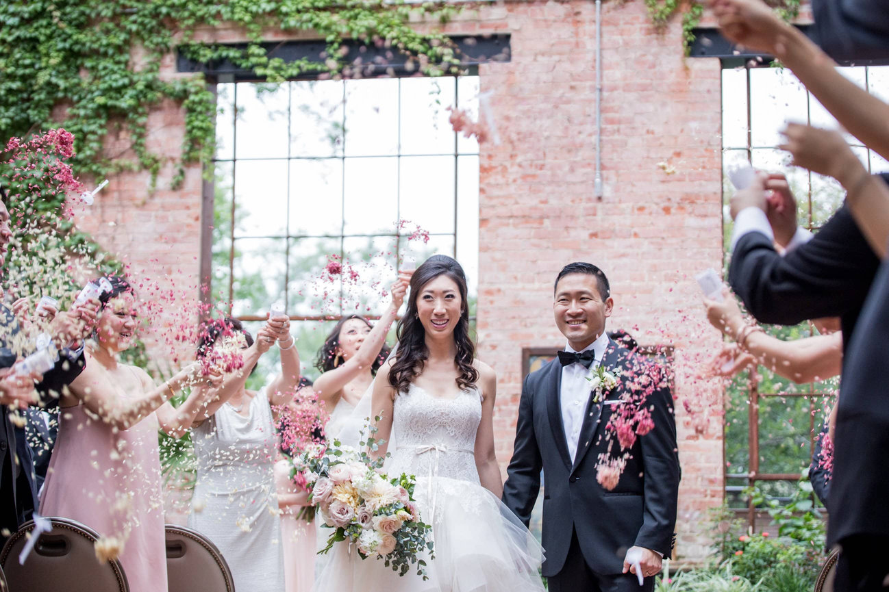 An exuberant wedding exit in San Antonio featuring an Asian bride and groom as flower petals are tossed; capturing the high-energy, editorial movement of a Texas celebration.