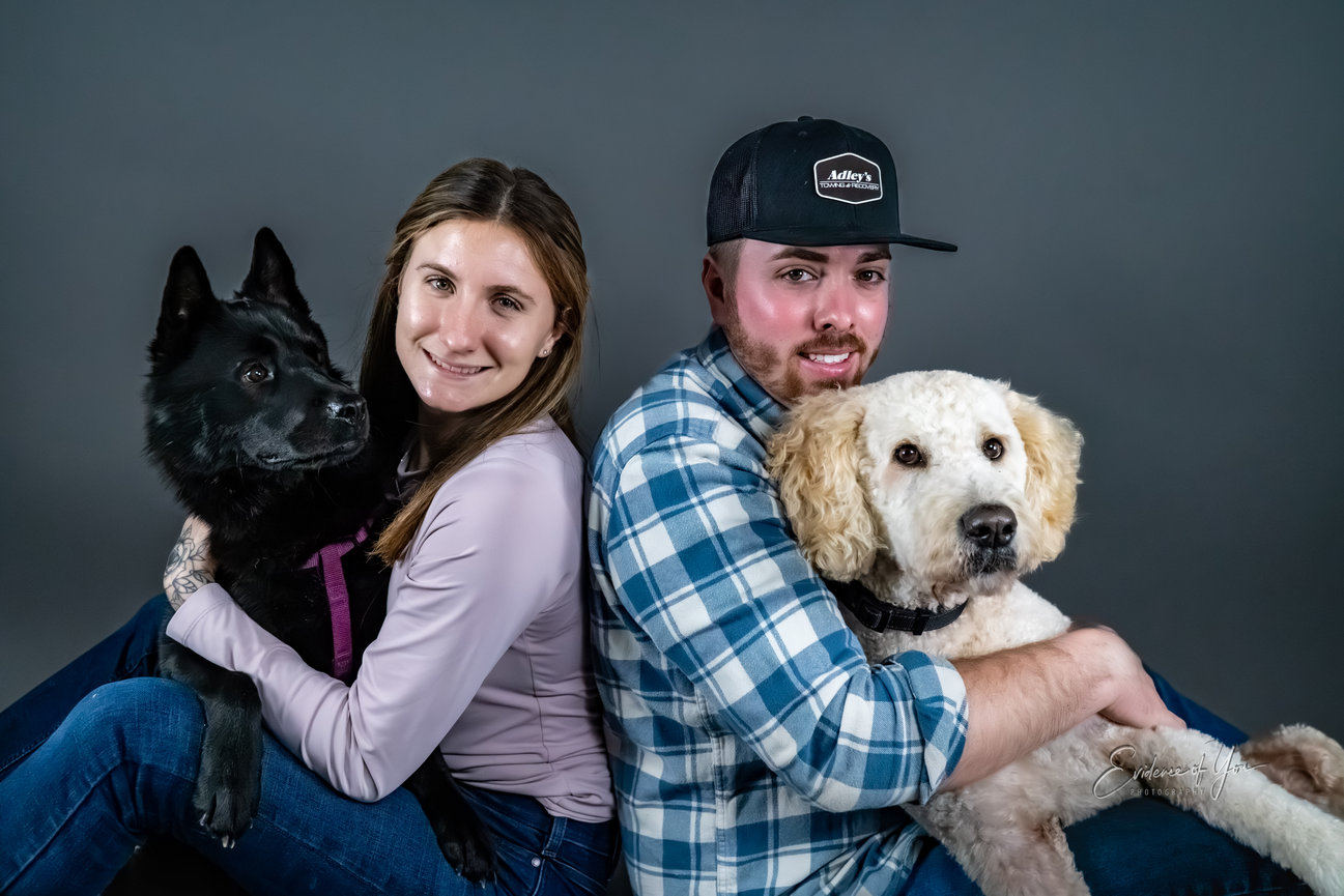 A couple seated back to back, each holding a dog against a gray background.