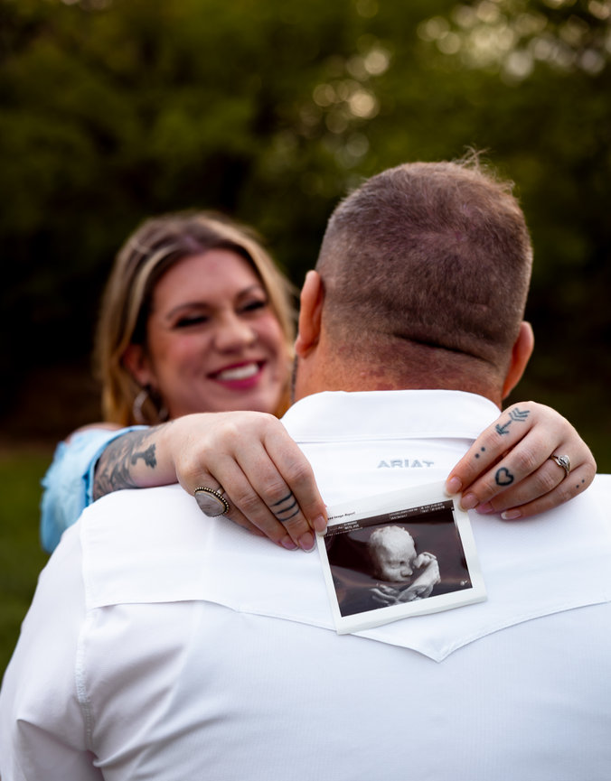 Woman smiling at a man holding an ultrasound photo, with trees blurred in the background.