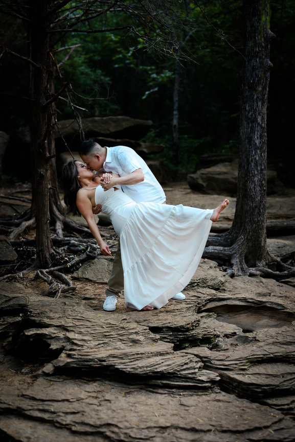 A couple elegantly dancing on a rocky forest path, surrounded by trees, the woman wearing a flowing white dress.