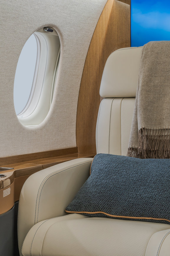 Luxurious private jet interior with a cream leather seat, blue pillow, beige throw blanket, and a window showing the sky.