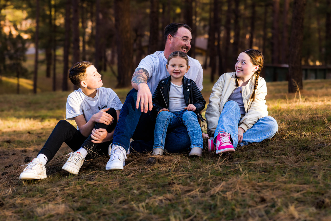 Father and three children enjoying an outdoor family portrait session in Ruidoso, NM, sitting together on the grass in a wooded area.