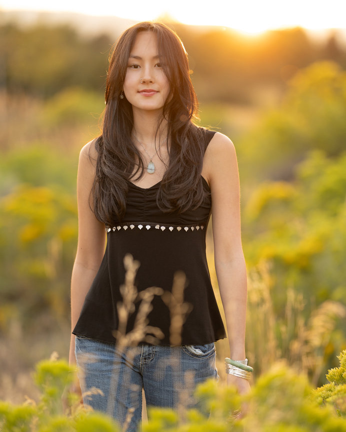 A woman with long hair in a black top and jeans stands in a grassy field during sunset.