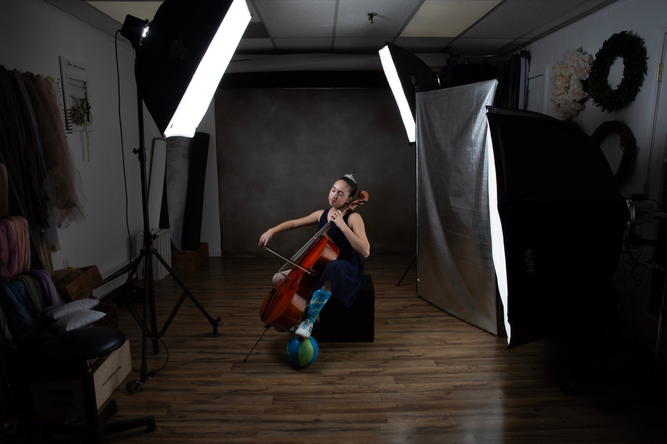 A teenage girl playing a cello in at Paula Swift photography photo studio with softbox lights and wooden floor.